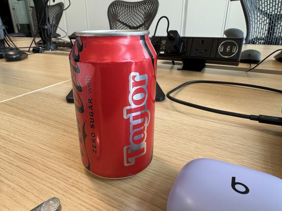 A red can of Coca-Cola Zero Sugar with the name "Taylor" on it sits on a wooden table. The background shows computer equipment and cables.