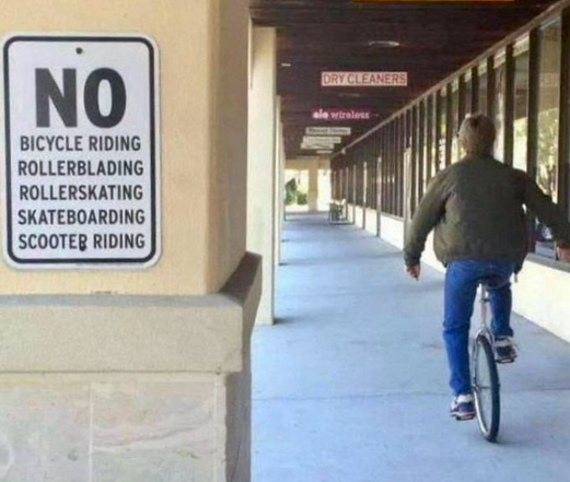 Someone on a unicycle rides past a sign prohibiting bicycles, rollerblades, roller skates, skateboards, and scooters.