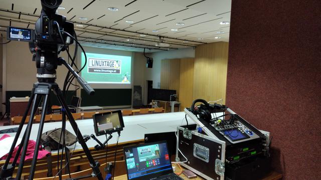 FOH Setup des c3voc hinten in einem Unihörsaal. Auf der Projektionsfläche das Kinuxtage Logo mit steyrer Tux. Von links nach rechts: Kamera auf Stativ, Vorschaumonitor, Mixerlaptop, Audiomischpult mit Funkempfängern.