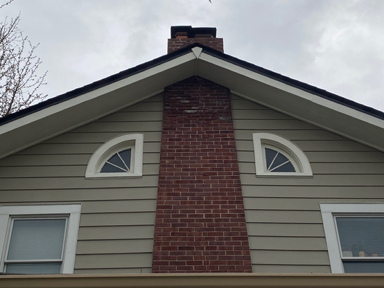 Color image of a residential home, windows bracketing the chimney. House is a 1920s balloon-frame colonial, lap sided in light green wood, attic windows bracketing both sides of the red brick chimney, windows below on the second floor likewise bracketing.