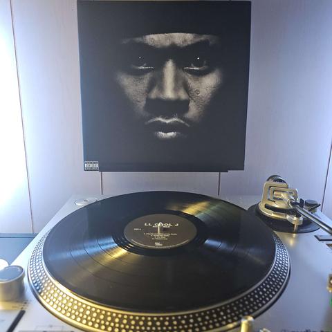 A vinyl record sits on a turntable. Behind the turntable, a vinyl album outer sleeve is displayed. The front cover shows LL Cool J's face up close.