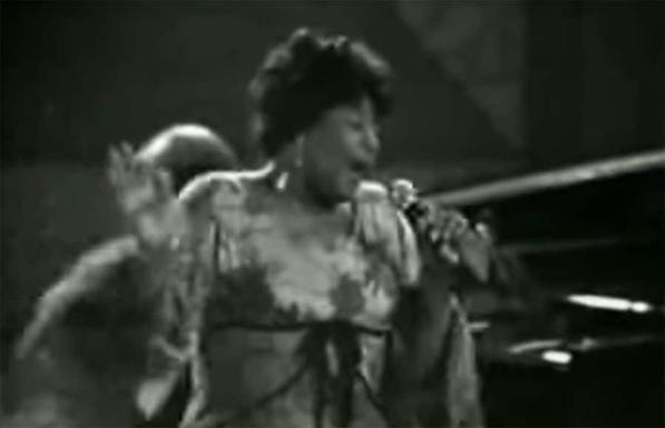 Black and white image of Ella Fitzgerald performing, mid-scat singing, with an expressive gesture and her hand near her ear as she sings at the Montreux Jazz Festival.
