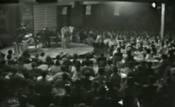 Black and white image of Ella Fitzgerald performing, mid-scat singing, with an expressive gesture and her hand near her ear as she sings at the Montreux Jazz Festival.