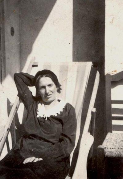 A woman lying on a deck chair in the harsh sunlight. She is wearing a dark outfit with a white collar.