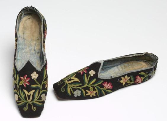 A pair of black wool slippers with square toes and a pointed vamp, decorated with red, yellow, white, and blue flowers and vines and leaves in several shades of green. The edges are bound in light blue silk, and the insoles appear to be the same silk, heavily worn.