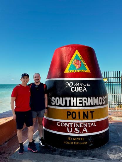 The Hao Travelers pose in front of the iconic Southernmost Point buoy in Key West, Florida. The large, brightly painted concrete marker stands against a backdrop of turquoise ocean and clear blue sky. The buoy is striped in red, black, yellow, and white and reads “90 Miles to Cuba – Southernmost Point – Continental U.S.A. – Key West FL, Home of the Sunset.” Nongchao wears a red shirt and dark shorts, and Mitchell wears a dark shirt and gray shorts, both looking relaxed and happy in the tropical setting.