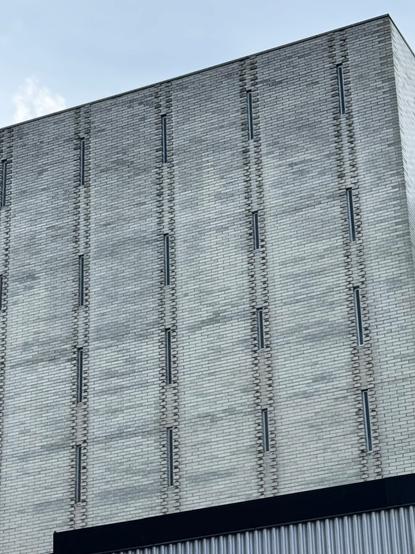 Photo of a section of a four-storey commercial building composed of grey bricks with very, very narrow windows.  The windows are so narrow they could almost be arrowslits.  The sky is pale blue.