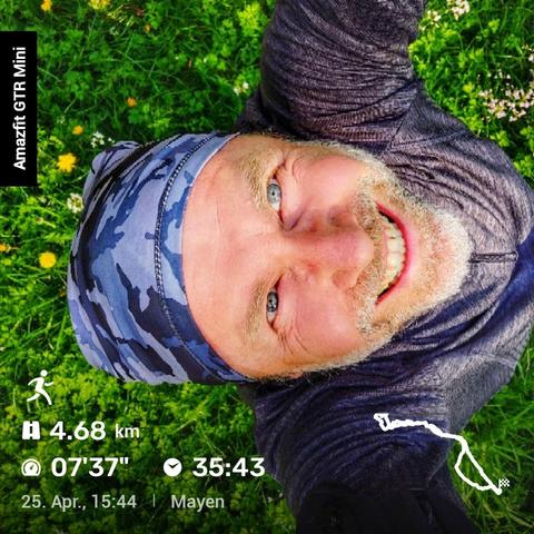 Das Bild zeigt eine Person in einem grauen Hoodie und einer Camouflage-Mütze, die auf einer Wiese mit gelben und weißen Blumen steht. Sie lächelt in die Kamera. Im eingeblendeten Text stehen Informationen wie ‚Amazfit GTR Mini‘, ‚4.68 km‘, ‚07'37''‘, ‚35:43‘ und das Datum ‚25. Apr., 15:44‘. Unten rechts ist eine kleine Karte mit der Laufroute zu sehen. Der Hintergrund vermittelt eine ruhige, naturverbundene Atmosphäre.