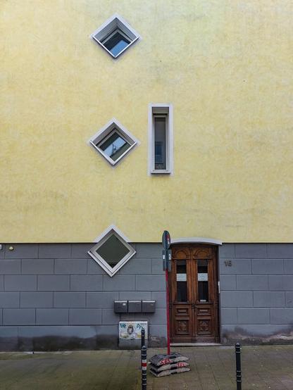 Ein gelbes Gebäude mit einer braunen Holztür und drei ungewöhnlich angeordneten Fenstern. Zwei der Fenster sind rautenförmig und eines ist rechteckig. Der untere Teil der Fassade ist grau gefliest. Vor der Tür stehen Sandsäcke und ein rot-weiß gestreifter Pfosten.