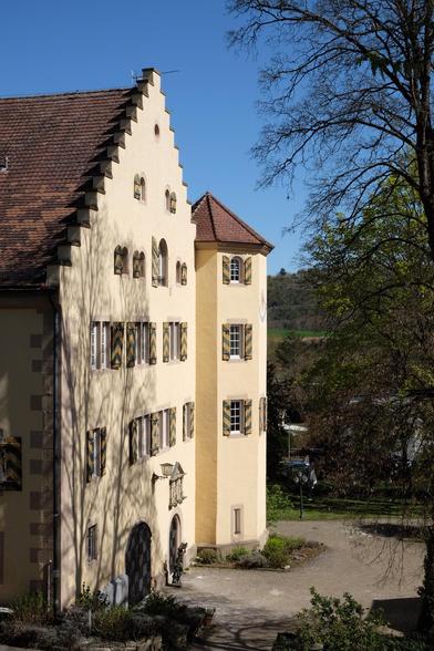 Hauptgebäude eines kleinen Schlosses mit hellgelben Mauern und gelb-grün gestreiften Fensterläden. Um das Gebäude führen Kieswege und ein kleiner Park.

Main building of a little castle with light yellow walls and yellow and green stripes on the shutters. Around the building are gravel paths and little park.