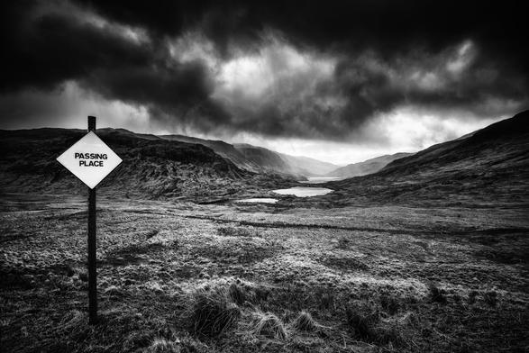 Schwarzweisse Landschaftsaufnahme auf der Hebrideninsel Mull in Schottland. Unter dramatischen Wolken sind Hügel und ein Tal mit drei Seen dargestellt; im Vordergrund links der Mitte ein rautenförmiges Schild mit der Aufschrift "Passing Place".