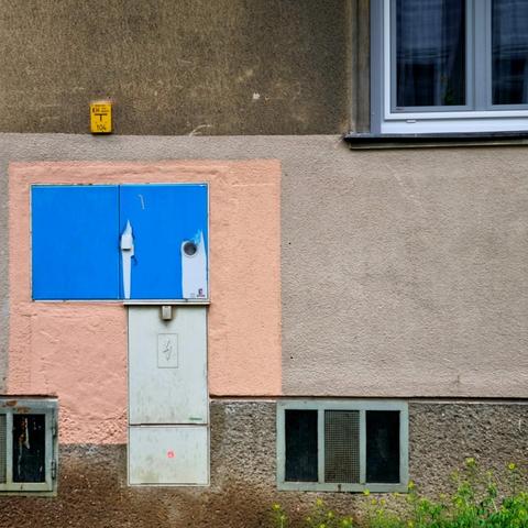 The square picture is a composition of partially overlapping rectangles (a bit Mondrian-like but without the bright colors) and shows a house wall. Horizontally, it is divided into three strips. A rough plastered wall with two cellar windows at the bottom. A dark façade with a window in the top. The left part of the middle strip shows a blue metal box (an electricity or gas connection) mounted on a salmon-colored wall. Below it is an upright box with a high voltage symbol. A small yellow box indicates the existence of gas.