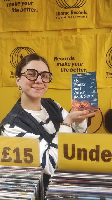 Rosie of Indie vinyl shop Thorne Records, holding up a paperback copy of My Family and Other Rock Stars by Tiffany Murray