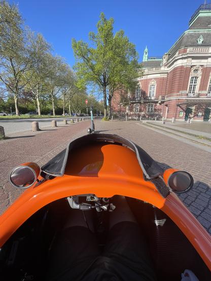 Endlich wieder mit dem Velomobil unterwegs, das Bild zeigt die Sicht des Fahrenden ein kajakartiger Einstieg ist am Rande zu sehen, davor erstreckt sich das orangene Vorderteil des Velomobils. Ich halte gerade um zu trinken und das vor der Musikhalle in Hamburg, welche in Hintergrund an der roten Backsteinfassade zu erkennen ist.