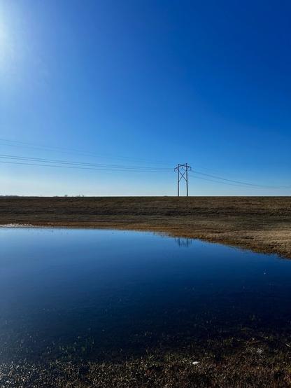 A large puddle of water next to a highway with bright blue skies overhead. A power pole is visible in the distance.