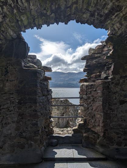 The view through a Y-shaped window in a stone wall. In the trunk of the Y, viewer can see a portion of the castle ruins, a sliver of the loch, and the hills on the opposite shore. Then the window opens up to show grey clouds and bright blue sky above.