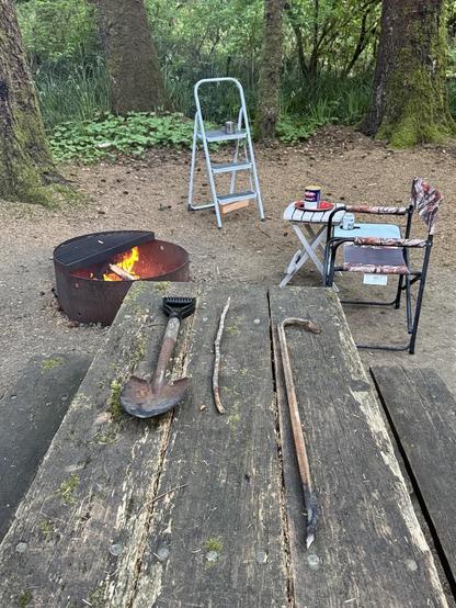 A wooden table in a forest setting holds a shovel, a stick, and a metal rod. In the background, there's a fire pit with flames, a folding chair, a small table, and a step ladder. Lush greenery surrounds the area.