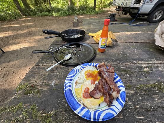 A campsite setup featuring a wooden table with a blue paper plate containing two eggs with yellow yolks and strips of bacon, garnished with ketchup. Nearby, there are two frying pans, a spoon, a bottle of hot sauce