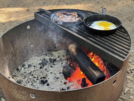A campfire cooking setup featuring two cast iron skillets. One skillet contains bacon sizzling, while the other has two frying eggs. The campfire is surrounded by a metal fire ring with glowing embers and ash.