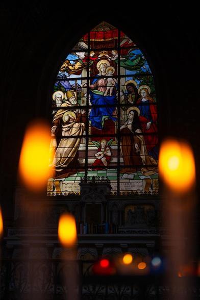 Ein mit katholischen Motiven bemaltes Kirchenfenster, eingerahmt von brennenden Kerzen im Vordergrund, die in der Unschärfe verschwinden.