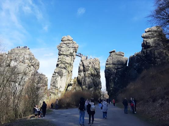 Das Bild zeigt die Externsteine, eine beeindruckende Landschaft mit hohen, felsigen Formationen, die sich majestätisch in den Himmel erheben. Der Himmel ist klar und blau mit wenigen weißen Wolken. Im Vordergrund befindet sich ein asphaltierter Weg, auf dem mehrere Menschen spazieren gehen und sich unterhalten. 

Text mit Unterstützung von @altbot