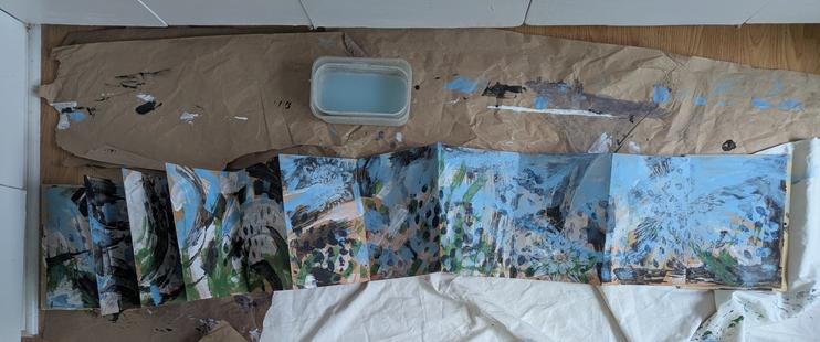 Accordion book, partially opened out, on brown paper and white shower curtain to protect floor. Portion shown covers about 1 x 5 feet. Framed around two edges by white kitchen cabinet fronts.

Large abstract strokes of black, white, muted light blue, indigo and "green earth" cover the paper randomly. Also, some indigo dots made with jumbo bubblewrap.