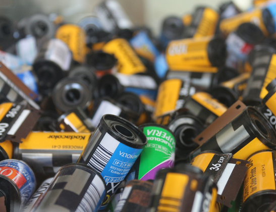 Long flat pile of 35mm film canisters. A few have film sticking out of them but most are probably empty. In the background they are out of focus and a section of white wall can be seen.