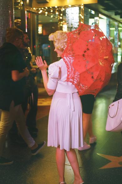 A Marilyn Monroe impersonator waves to a modern crowd passing by on Hollywood blvd, evening time.