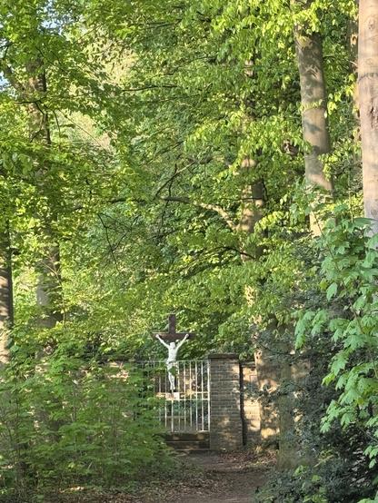 Im frischen Grün der Bäume sieht man eine Mauer mit eisernem Tor, weiter hinten Jesus am Kreuz