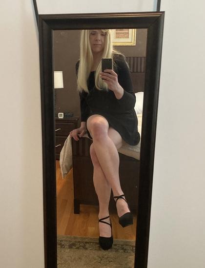 mirror selfie of blonde woman with thigh length black dress and black strap high heels sitting on the edge of a bed cross-legged