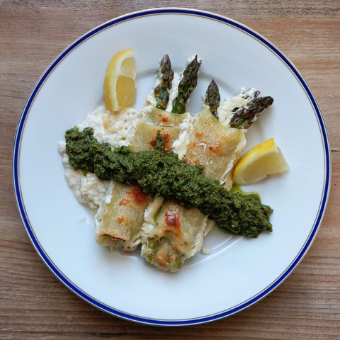 Auf einem Teller zwei mit grünem Spargel gefüllte Pasta-Rollen (Canneloni) mit grünem Pesto + 2 Zitronenspalten und etwas von der Béchamel