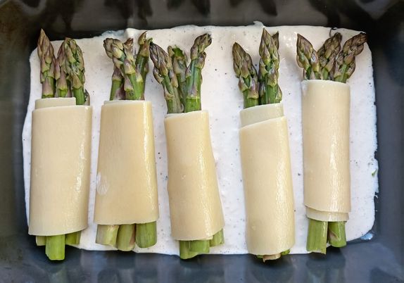 in einer Auflaufform ein Teil der Sauce, darauf liegen 5 jeweils mit 3 Stangen grünem Spargel gefüllte Pasta-Rollen (Canneloni)