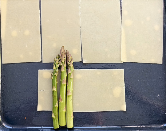 Auf einem Backblech liegen 5 Lasagneplatten. Auf einer liegen 3 Stangen grüner Spargel, die oben und unten über die Platte herausragen.