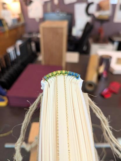 The top edge of a book showing the handsewn endband. The endband is a gradient of colors including blue, green, and gold.