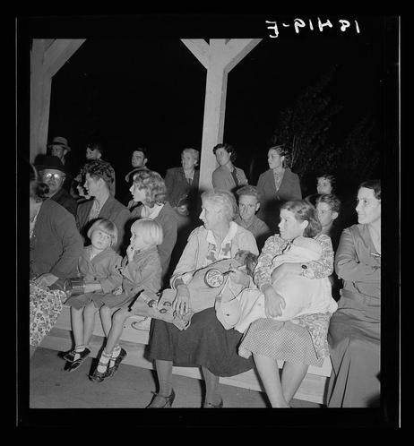 The image is a black and white photograph depicting a group of people seated outdoors at night. The setting appears to be informal, possibly part of an outdoor event or gathering. There are men, women, and children present in the scene.

Several individuals seem engaged with one another, while others appear more isolated within their own company. A few young girls hold stuffed animals, suggesting a casual, perhaps family-oriented atmosphere. Clothing styles hint at mid-20th-century fashion norms: suits for adult males, dresses and skirts for females, and overcoats or lighter garments suitable for cooler nighttime temperatures.

Visible text on the image includes what appears to be "E-PHIPI" in Hebrew characters followed by "F-PHPI," which could indicate a date format (10-45) or possibly reference another location. The context provided suggests that this photograph captures mothers observing an event from afar, likely reflecting their roles within the community and responsibilities as caretakers.

The overall mood conveyed is one of quiet observation, with the subjects' expressions ranging from contemplative to engaged. This snapshot seems to document a social moment in time for individuals who may have been part of migrant or labor camps during that era, based on additional information provided about its origin.