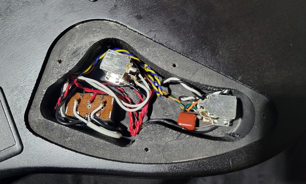 Control cavity detail of an electric guitar. It features 3 main components, a box switch, and 2 push/pull potentiometers. On the left is the switch, the volume pot is above it just off center to the right, and the tone control is seen on the far right. An orange,  square-ish .022 micro farad capacitor is seen coming off the tone control. A series of colored, twisted pair wires connects them all together into a circuit. The back of the guitar has a matte black finish. The background is black fabric.