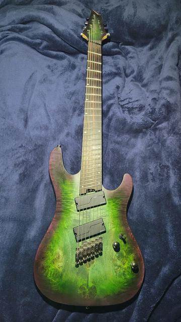 Image of a 7 string, multiscale (fanned fret) Cort electric guitar. The body shape is very traditional, and features a burl top. It is green, with an outline of purple around the edges. The pickups and frets are all angled, due to the nature of a multiscale instrument. The electronics feature 2 knobs and one switch. One volume, one tone, and one pickup selector switch. 7 individual string bridges truncate the string terminations.
