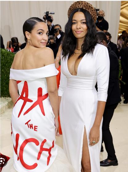 Das Bild zeigt zwei Frauen auf einem roten Teppich, vermutlich bei einer prominenten Veranstaltung wie der Met Gala. Beide tragen elegante weiße Kleider. Die Frau links trägt ein schulterfreies Kleid, auf dessen Rückseite in großen roten Buchstaben der Schriftzug „TAX THE RICH“ steht – eine politische Botschaft („Besteuert die Reichen“). Ihr Haar ist glatt zurückgekämmt und zu einem Knoten gestylt. Die Frau rechts trägt ein tief ausgeschnittenes Kleid mit einem Beinschlitz und einer auffälligen, braun gemusterten Kopfbedeckung (vermutlich im Leoparden-Look). Sie hat lange, lockige Haare und ein selbstbewusstes Lächeln. 🖖