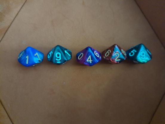 The image shows five polyhedral dice arranged in a row on a brown surface. The dice are of different colors and have a metallic finish with a shiny, reflective surface. The first die is blue with the number 1 facing up. The second die is teal with the number 9 facing up. The third die is purple with the number 4 facing up. The fourth die is a mix of brown and red with the number 2 facing up. The fifth die is teal with the number 5 facing up. Each die has white numbers on a colored background, making them stand out. The dice are slightly angled, giving a three-dimensional perspective to the image.