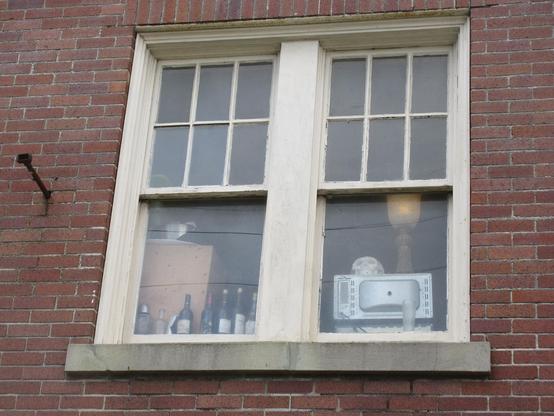 View to upstairs window in an older brick building.  Seen in the window are several upright wine bottles, the back of a microwave oven, and a rubber or plastic model of a human skull placed to look out the window.