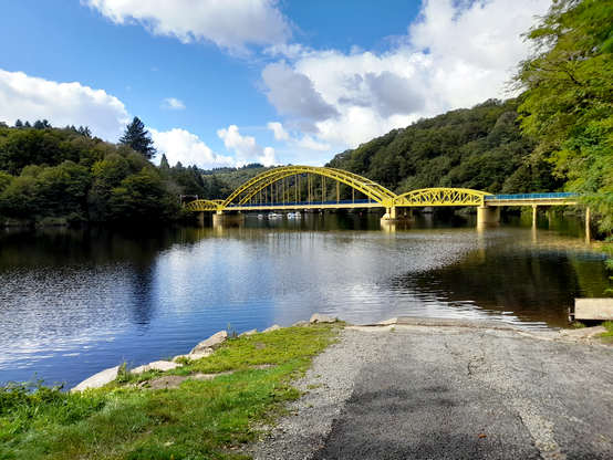 A road ending at the edge of a river bank. Further away is a large yellow bridge across the water.