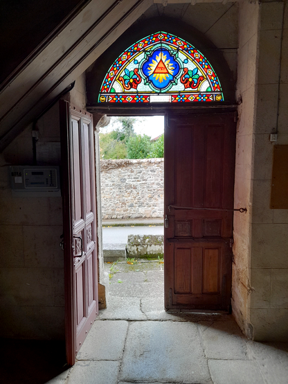 A picture of a church door taken from inside. One half of the door is open and above it is a stained-glass window in red, green, yellow and blue with flower and a triangle with a text inside that appears to be hebrew.