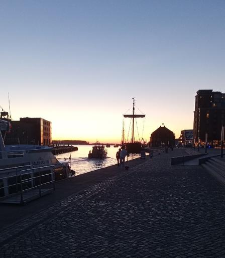 View of the outer end of the old harbour basin in Wismar with the police patrol vessel "Walfisch" passing the moored "Wissemara", a replica of a medieval Hanseatic League kogg ship. Brick storehouses from the 19th and 20th centuries, now repurposed as holiday apartments, to the right and ine of the oh so common "entrepreneurial centres at the waterfront" to the left. In mid distance a young woman standing next to the basin, posing against the yellow evening sky for a friend who is taking some pictures of her.