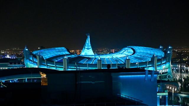 A curvy designed stadium at night, with a big sculpture of a horse head hovering over it. Everything is illuminated by blue lighting.