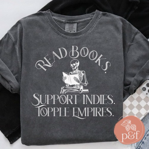 Dark grey t-shirt with the words: “Read Books. Support Indies. Topple Empires.” An image of a skeleton reading is in the middle of the shirt.