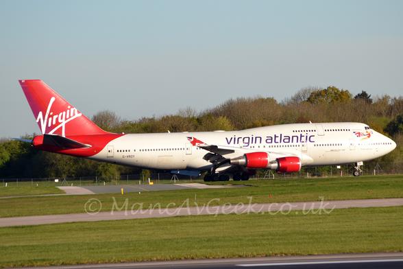Side view of a white, 4 engined jet airliner  taxiing from left to right with flaps extended from the rear of the wings, suggesting it has just landed.
The plane has purple "Virgin Atlantic" titles on the upper mid-fuselage, with an image of a woman wearing a red mini-dress flying through the air and trailing a Union Flag positioned under the cockpit windows on the forward fuselage.
The tail is a bright metallic red, with seemingly hand-drawn "Virgin" titles.
The engine pods slung under the wings are the same metallic red.
The squared-off up-turned wingtips have a waving modified Union Flag.
Green grass fills the foreground, with trees dominating the background, under grey-blue skies.