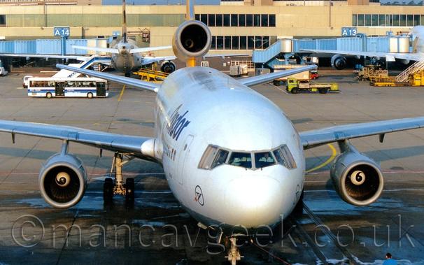 Almost head-on view of a white, 3 engined jet airliner facing slightly off to the right of the camera, just a few metres away from it's parking spot.
There are blue "Condor" titles on the upper forward fuselage, and the blue outline of a circle with a stylised blue flying bird in the middle, on the sides of the nose under the cockpit side windows.
The tail and the centre engine pod are a bright yellow.
Grey concrete apron fills the foreground and most of the background, marked with yellow lines for the planes to follow and white lines for other vehicles.
A pale, sandy brown terminal building stretches across the frame, with blue airbridges hanging off the side, some attached to white jet airliners.
Lime green trucks and white buses  are crossing the frame in the background, with more vehicles parked in the shadows of the other planes.