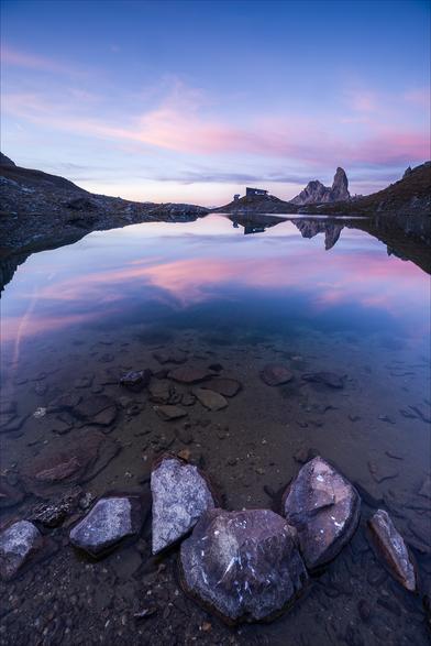 Photo d'un paysage de montagne au lever du jour, au format portrait.
Au premier plan, des cailloux baignent dans un lac, probablement près du bord vu la faible profondeur.
Tout au bout du lac se dressent un refuge et une montagne en forme de canine. Le ciel est bleu avec de fin nuages roses qui se reflètent dans le lac.
-
Portrait format photo of a mountain landscape at sunrise.
In the foreground, pebbles bathe in a lake, probably close to the shore given the shallowness of the water.
At the far end of the lake stands a refuge and a canine-shaped mountain. The sky is blue, with fine pink clouds reflected in the lake.