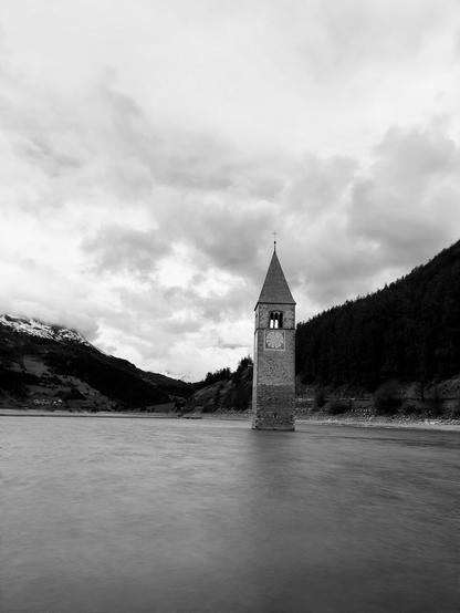 A black and white image of a submerged bell tower in a lake, surrounded by mountains and cloudy skies. The clock face on the tower is visible, highlighting its historical significance.