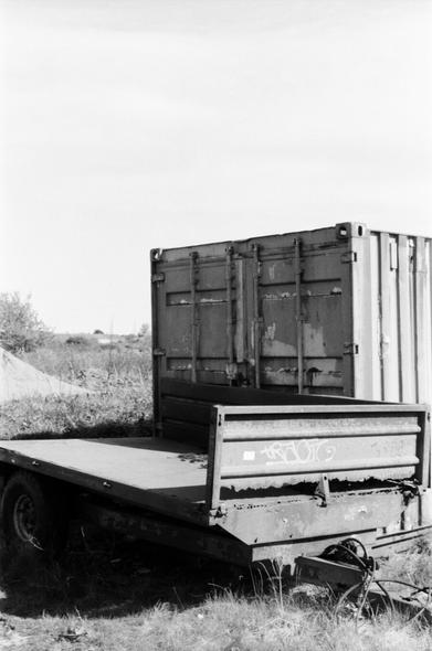 Black and white photo of a shipping container with a rusted, broken trailer in front of it
